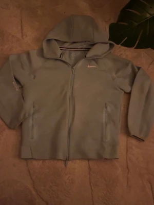 Grå NOCTA Nike hoodie med dragkedja - Snygg grå hoodie från Nike NOCTA med hel dragkedja och två fickor framtill. Hoodien har en klassisk huva och diskret Nike-logga på bröstet samt NOCTA-tryck på ärmen. Perfekt för en streetwear-look och tillverkad i mjukt material.