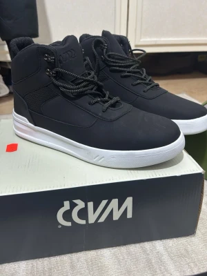 Svarta höga sneakers från WAVE - Stilrena svarta sneakers från WAVE med högt skaft och vit sula. Skorna har svarta snören med vita detaljer och vadderad insida för extra komfort. Materialet är en mix av syntet och textil, vilket ger en modern look. Perfekta för dig som gillar streetwear.
