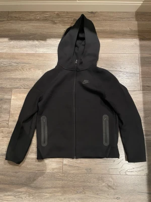 Nike tech - Svart hoodie från Nike med hel dragkedja framtill och två fickor med dragkedja. Klassisk design med huva och diskret Nike-logga på bröstet. Perfekt för en avslappnad och sportig stil.