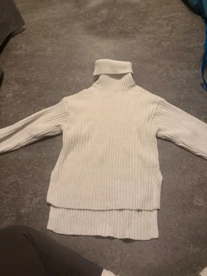 Vit ribbstickad polotröja - Mysig vit ribbstickad polotröja med hög krage och långa ärmar. Tröjan har en avslappnad passform och slitsar i sidorna, vilket ger en trendig look. Perfekt för lager på lager och enkel att matcha med olika outfits.