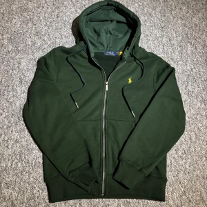 Mörkgrön hoodie från Polo Ralph Lauren - Mörkgrön hoodie med dragkedja från Polo Ralph Lauren. Klassisk design med huva, snörning och gul broderad logga på bröstet. Mjukt material och ribbade muddar för en skön passform. Perfekt för en avslappnad och stilren look. Storleken är lite mindre så egentligen M men passar för S. Hör av dig vid frågor om pris eller liknande 😊