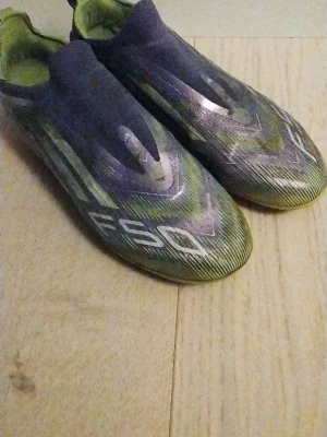 Adidas F50 Laceless Elite Radiant Blaze barn fotbollsskor lila/vit/Lucid/lemon - Säljer ett par Adidas F50 fotbollsskor i grön och lila med silvriga detaljer. Skorna har en strömlinjeformad design utan snörning och mönstrad ovandel. Yttersulan är gulvit med fasta dobbar, perfekt för spel på gräsplan.