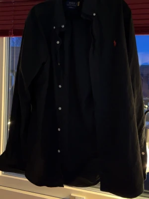 Svart skjorta från Polo Ralph Lauren - Klassisk svart skjorta från Polo Ralph Lauren med vit knappstängning och röd broderad logga på bröstet. Skjortan har lång ärm och en stilren, tidlös design som passar till många olika outfits.