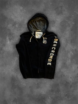 Svart hoodie från Abercrombie - Snygg svart hoodie från Abercrombie & Fitch med dragkedja och stor huva. Grått innerfoder i huvan och vita snören. Stor logga i vitt längs ena ärmen och tryckt emblem på bröstet. Perfekt för en chill och avslappnad stil.