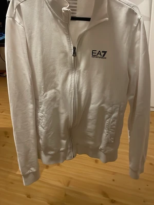 Vit ziptröja från EA7 Emporio Armani - Snygg vit ziptröja från EA7 Emporio Armani med dragkedja framtill och två fickor. Tröjan har hög krage och diskret logga på bröstet. Perfekt till chill eller streetwear-stil. Tillverkad i mjukt material för skön känsla.