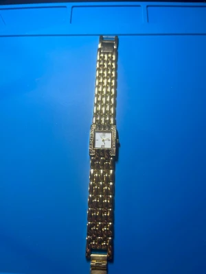 Guldfärgad fyrkantig klocka - Snygg guldfärgad klocka med fyrkantig urtavla och länkarmband. Urtavlan är vit med diskreta markeringar och ramen är dekorerad med små stenar för extra bling. Perfekt accessoar för dig som vill sticka ut. Behövs nya batterier 