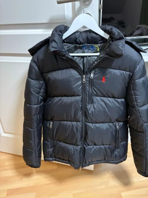 Svart pufferjacka från Polo Ralph Lauren - Snygg svart pufferjacka från Polo Ralph Lauren med röd logga på bröstet. Jackan har dragkedja framtill, två sidofickor med dragkedja och en bröstficka. Fylld för extra värme och har en hög krage samt avtagbar huva.