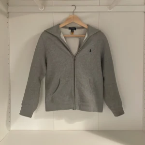  Polo Ralph Lauren zip up - Grå hoodie från Polo Ralph Lauren med dragkedja framtill och klassisk broderad logga på bröstet. Tröjan har huva, två fickor och är tillverkad i mjuk bomullsblandning. Perfekt för chill dagar eller när du vill ha en clean och enkel look.
