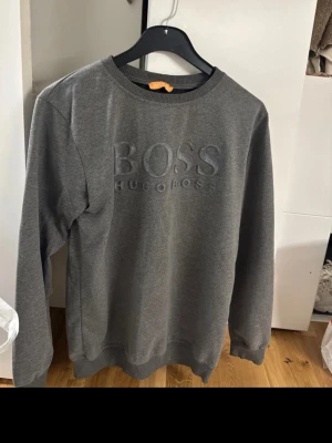 Grå sweatshirt från Hugo Boss - Säljer en stilren grå sweatshirt från Hugo Boss med broderad logga framtill. Tröjan har rund hals, ribbade muddar och långa ärmar. Perfekt för dig som gillar klassisk design med en modern touch.