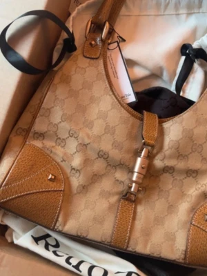 Beige axelremsväska från Gucci - Snygg beige axelremsväska från Gucci med klassiskt GG-mönster och bruna läderdetaljer. Väskan har en unik metallspänne framtill och justerbar axelrem. Perfekt storlek för det viktigaste och ger en lyxig touch till din outfit. Inköpt från A retro tale och i mycket fint skick! Har artikeln publicerad på Vinted också, först till kvarn. 
