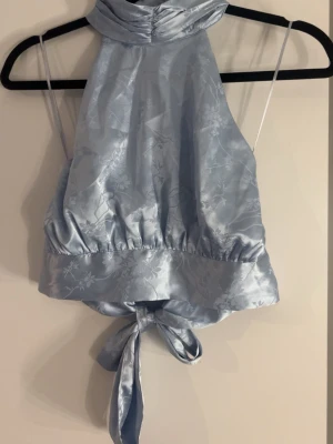 Ljusblå halterneck topp med knyt - Supersnygg ljusblå halterneck topp i glansigt satintyg med subtilt blommönster. Toppen har hög hals och knytning i ryggen för en trendig look. Perfekt att styla med jeans eller kjol för en cool vibe.