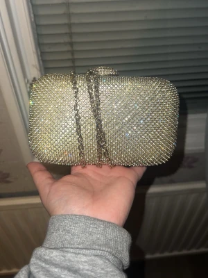 Glittrig guldig kuvertväska med kedja - Säljer en superglittrig kuvertväska i guld med massor av små strass-stenar och en snygg guldfärgad kedja. Väskan är hård och har en rektangulär form med metalliska detaljer på sidorna. Perfekt för dig som vill sticka ut och älskar bling!