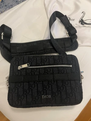 Svart Dior axelväska med monogram - Säljer en svart axelväska. Väskan har silverfärgade dragkedjor, justerbar axelrem och en praktisk ytterficka. Kommer med original dustbag och exklusiv presentbox med blått och guldmönster.