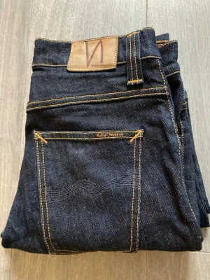 Mörkblå jeans från Nudie Jeans - Snygga mörkblå jeans från Nudie Jeans med klassisk femficksdesign och kontrastsömmar. Grim trim. Jeansen har rak passform och är tillverkade i kraftig denim med läderpatch bak i midjan. Perfekta för en stilren och avslappnad look. Nya, knappt använda