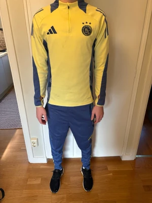 Ajax träningsoverall gul/blå Adidas - Ajax träningsoverall från Adidas i gult och blått. Överdel med halv dragkedja, klubbmärke och Adidas-logga på bröstet samt tre stjärnor. Matchande blå byxor med klubbmärke och gula detaljer. Perfekt för fotbollsträning eller chill. Personen i bilden är 184 cm