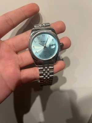 Silverfärgad klocka från Sunkta - Stilren klocka från Sunkta med silverfärgat armband i metall och ljusblå urtavla. Klockan har arabiska siffror, klassisk rund form och datumfönster. Perfekt accessoar för dig som gillar en clean och modern look. 