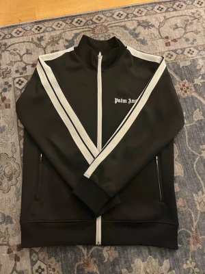Palm Angels svart track jacket - Svart track jacket från Palm Angels med vita ränder längs ärmarna och dragkedja framtill. Jackan har ståkrage, två fickor med dragkedja och logga på bröstet. Perfekt för en sportig och trendig look.