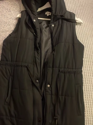 Svart lång dunväst med huva från Bubbleroom  - Säljer en svart lång dunväst från Bubbleroom med huva och dragsko i midjan. Västen har två fickor framtill, dragkedja och tryckknappar. Perfekt för lager på lager och riktigt snygg till höst- och vinteroutfits.