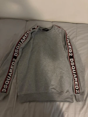 Grå sweatshirt från Dsquared2 - Snygg grå sweatshirt från Dsquared2 med svarta och röda logoband längs båda ärmarna. Tröjan har rund halsringning och ribbade muddar vid ärmslut och nederkant. Perfekt för en avslappnad och trendig stil.