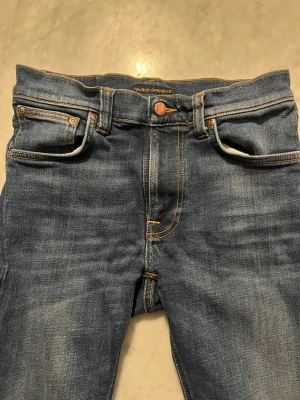 Blå jeans från Nudie Jeans Co - Säljer ett par klassiska blå jeans från Nudie Jeans Co med fem fickor, orangea sömmar och läderpatch med logga bak. Jeansen har en snygg tvättad look och raka ben. Perfekta för dig som gillar stilrena och tidlösa jeans. Jeansen är i väldigt bra skick. Kan gå ner i pris om affären är smidig.