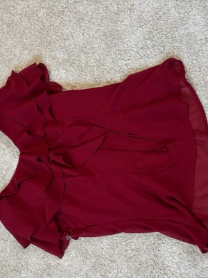 Vinröd blus med volanger - Snygg vinröd blus i ett tunt, transparent tyg med volangdetaljer längs axlar och halsringning. Blusen har korta ärmar och ett knytband framtill som ger en feminin touch. Perfekt att styla med jeans eller kjol för en trendig look.