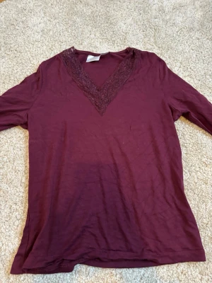 Vinröd långärmad topp med spets - Vinröd långärmad topp med v-ringning dekorerad med spetsdetaljer. Tillverkad i mjukt tyg, perfekt för en stilren och enkel look. Passar till många olika outfits och har en smickrande passform.