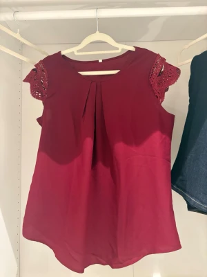 Vinröd topp med spetsdetaljer - Snygg vinröd topp med rund halsringning och spetsdetaljer på ärmarna. Luftig passform och korta ärmar med vacker virkad spets som ger en romantisk touch. Perfekt att styla med jeans eller kjol.