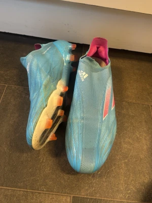 Blåa Adidas fotbollsskor utan snörning - Säljer ett par blåa Adidas fotbollsskor med rosa och vita detaljer. Skorna har en strömlinjeformad design utan snörning och är tillverkade i syntetmaterial. Yttersulan har orange dobbar för bra grepp på planen. Perfekta för dig som vill sticka ut på matchen!  Mitt pris 1000, använda lite mer än en halvsäsong!