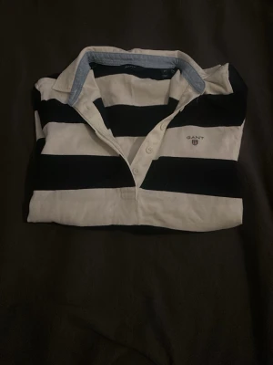 Randig långärmad skjorta från Gant - Snygg långärmad skjorta från Gant med breda svarta och vita ränder. Klassisk krage med knappar och diskret logga på bröstet. Tillverkad i mjuk bomull, perfekt för en avslappnad men stilren look.