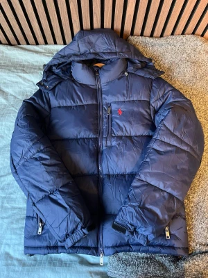 Mörkblå pufferjacka från Polo Ralph Lauren - Snygg mörkblå pufferjacka från Polo Ralph Lauren med röd logga på bröstet. Jackan har huva, dragkedjor på fickorna och bröstficka samt quiltad design. Perfekt för kalla dagar och riktigt skön att bära. Den är använd två gånger, priset kan diskuteras vid snabb affär ✅