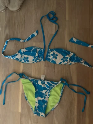 Blåvit blommig bikini med knyt - Snygg bikini med blått och vitt blommönster. Överdelen har knyt både i nacken och ryggen, och underdelen har knyt i sidorna samt limegröna detaljer på insidan. Perfekt för stranden eller poolen!