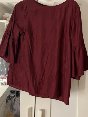 Vinröd blus med vida ärmar från Lindex - Snygg vinröd blus från Lindex med vida, trekvartsärmar och rund halsringning. Tillverkad i ett mjukt, vävt tyg som ger ett elegant fall. Perfekt att styla med jeans eller kjol för en trendig look.