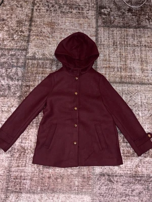 Vinröd kappa med huva - Stilren vinröd kappa med huva och knappar framtill. Kappan har två fickor och en rak passform. Aldrig använd! Pris kan diskuteras 
