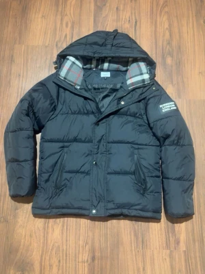 Svart dunjacka från Burberry - Snygg svart dunjacka från Burberry med huva och klassiskt rutigt innerfoder vid kragen. Jackan har två snedställda fickor framtill och en tydlig Burberry London England-logga på ärmen. Perfekt för kalla vinterdagar.