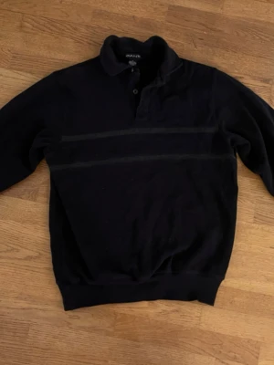 Svart tröja med krage från Gant - Klassisk svart tröja från Gant med krage och knappar vid halsen. Tröjan har diskreta horisontella ränder över bröstet och ribbade muddar vid ärmslut och nederkant. Perfekt för en stilren och avslappnad look.