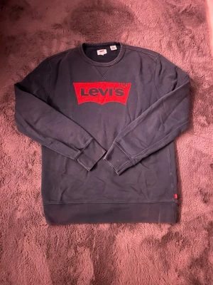 Svart sweatshirt från Levi's med logga - Cool svart sweatshirt från Levi's med den klassiska röda loggan framtill. Tröjan har rund halsringning, långa ärmar och ribbade muddar. Perfekt för en avslappnad och stilren look.