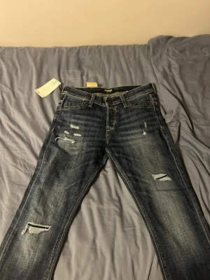 Slim Glenn blå slitna jeans Jack & Jones - Säljer ett par Slim Glenn jeans från Jack & Jones i mörkblå tvätt med slitningar och trasiga detaljer på benen. Jeansen har fem fickor sopm klassiska jeans och är gjorda i bomull med lite stretch för skön passform. Snygg slim fit-modell som ger en modern look. Ny pris 800 säljer för 600