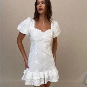 Vit kort klänning med puffärm - Superfin vit kort klänning med puffärmar, broderidetaljer och volang nertill. Klänningen har broderade blommor, fyrkantig urringning och en jättefin öppen rygg med söt rosett. Perfekt för studenten! Helt ny med prislapp!