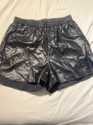 Svarta shorts i skinnimitation - Snygga svarta shorts i skinnimitation med elastisk midja och en bakficka. De har en loose passform och ger en cool look till din outfit. Perfekta för dig som vill sticka ut lite extra.