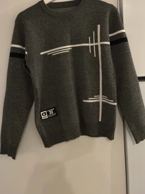 Grå stickad tröja med grafiskt mönster - Snygg grå stickad tröja med vita och svarta grafiska linjer på framsidan och armarna. Tröjan har rund halsringning, ribbade muddar och en patch med text nertill. Perfekt för dig som gillar modern och stilren design.