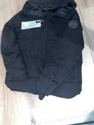 Svart dunjacka från Canada Goose - Snygg svart dunjacka från Canada Goose med huva och diskret logotyp på ärmen. Jackan har dragkedja framtill och två sidofickor. Tillverkad i slitstarkt material och fodrad med dun för extra värme. Perfekt för kalla vinterdagar.