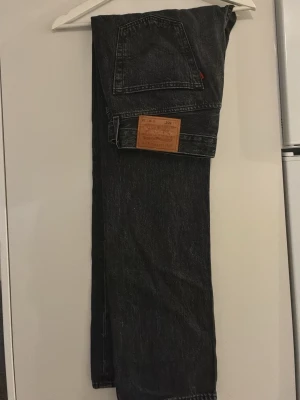 Svarta Levi's 501 jeans raka ben - Klassiska svarta Levi's 501 jeans med raka ben och femficksmodell. Jeansen har en brun läderpatch bak i midjan och synliga kontrastsömmar. Tillverkade i 100% bomull och har en tidlös, avslappnad passform.