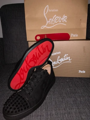 Svarta Christian Louboutin sneakers - Snygga svarta sneakers från Christian Louboutin med ikoniska röda sulor och svarta nitar på tån. Skorna har snörning och är tillverkade i mocka och läder. 