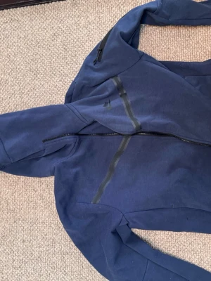 Blå hoodie från Nike med dragkedja - Snygg blå hoodie från Nike med hel dragkedja framtill och huva. Hoodien har svarta detaljer och en diskret Nike-logga på bröstet. Tillverkad i mjukt material som passar perfekt för en avslappnad stil.