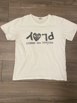 Vit Comme des Garçons Play t-shirt - Vit t-shirt från Comme des Garçons Play med det ikoniska hjärtlogotyp-trycket och texten 'PLAY' i svart på bröstet. Klassisk rund hals och korta ärmar. Tillverkad i mjuk bomull för en skön och avslappnad känsla. Passar M