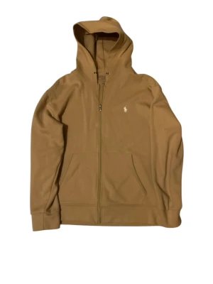 Polo Ralph Lauren Zip Hoodie - Snygg beige hoodie med dragkedja från Polo Ralph Lauren. Tröjan har huva med dragsko, två fickor framtill och den klassiska broderade loggan på bröstet. Perfekt för en avslappnad och stilren look.