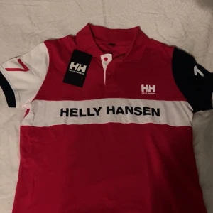 Röd och vit pikétröja Helly Hansen - Snygg pikétröja från Helly Hansen i rött, vitt och svart med klassisk krage och knappar framtill. Stor logga på bröstet och tryck på ena ärmen. Perfekt för en sportig och stilren look.
