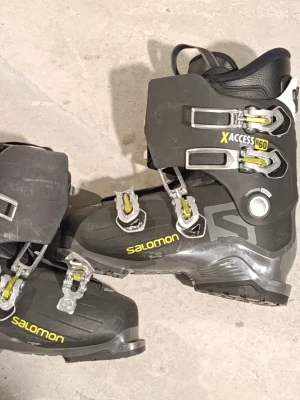 Salomon X Access 60 pjäxor  - Säljer ett par svarta Salomon X Access 60 pjäxor med gula detaljer och metallspännen. Skalet är robust i plast och designen är sportig med justerbara spännen för perfekt passform. Perfekta för dig som vill ha stabilitet och komfort i skidbacken.