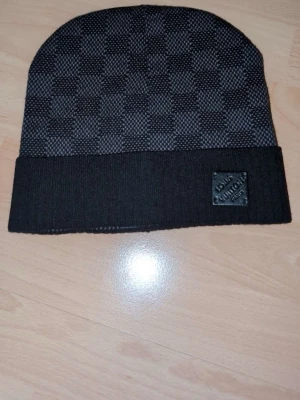 Louis Vuitton svart och grå mössa - Snygg svart och grå mössa från Louis Vuitton med klassiskt rutmönster och vikt kant. Märkeslapp i läder framtill och diskret LV-detalj. Perfekt accessoar för dig som vill ha något exklusivt och stilrent till hösten och vintern.