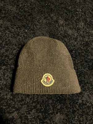 Grå ribbstickad Moncler mössa - Säljer en grå ribbstickad mössa från Moncler med uppvikt kant och det klassiska Moncler-logotypen framtill. Mössan är tillverkad i mjukt material och har en enkel, stilren design som passar perfekt till höst och vinter.